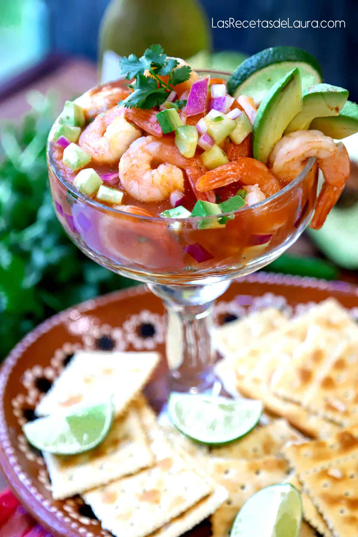 A glass of Mexican shrimp cocktail on a plate with hot sauce in the background. 